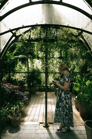 A lady in a flowery dress standing in a glass house full of plants and watching her telephone