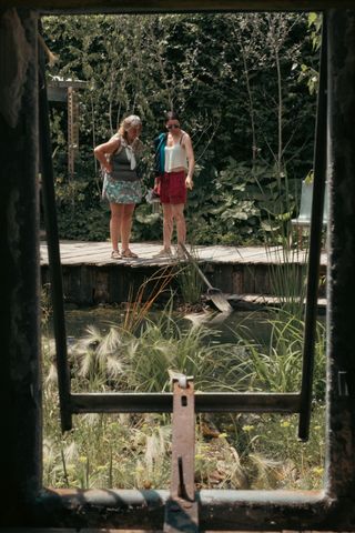 Seeing through a frame two woman are looking down on a pond with one of them pointing something in it