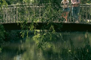 Two women under a weeping willow leaning on a bridge over the water