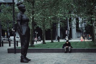 A woman waiting, sitting and watching her phone next to a tall black statue of a woman standing and waiting in the city